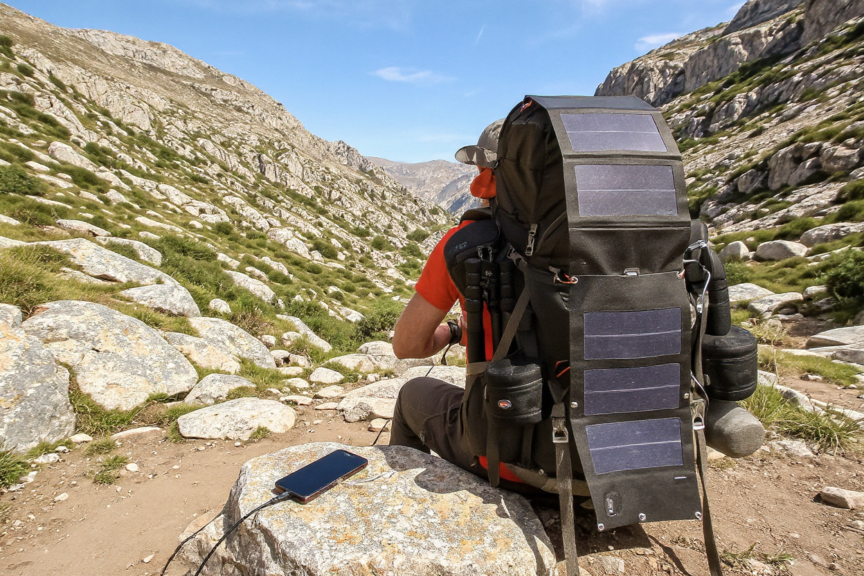 Chargeur solaire portable déplié sur un sac à dos, rechargeant un smartphone au soleil sur un sentier.