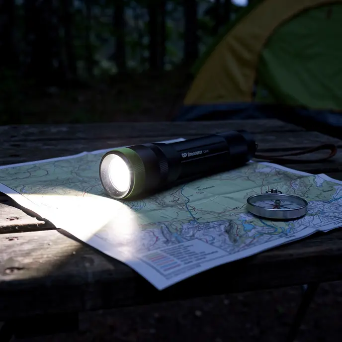 Lampe torche tactique posée sur une table en bois avec carte de camping et boussole au crépuscule