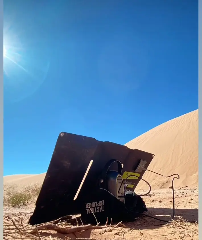 Tablette/écran solaire portable posé dans le sable, relié à une batterie externe et câbles, sous un ciel bleu en plein désert.