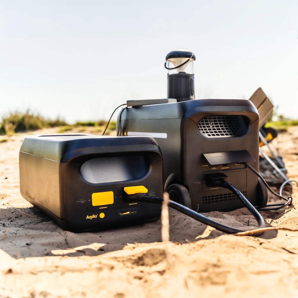 Chargeur/stockage énergétique portable sur plage: blocs d’alimentation noirs avec poignées et câbles connectés, disque solaire en arrière-plan.