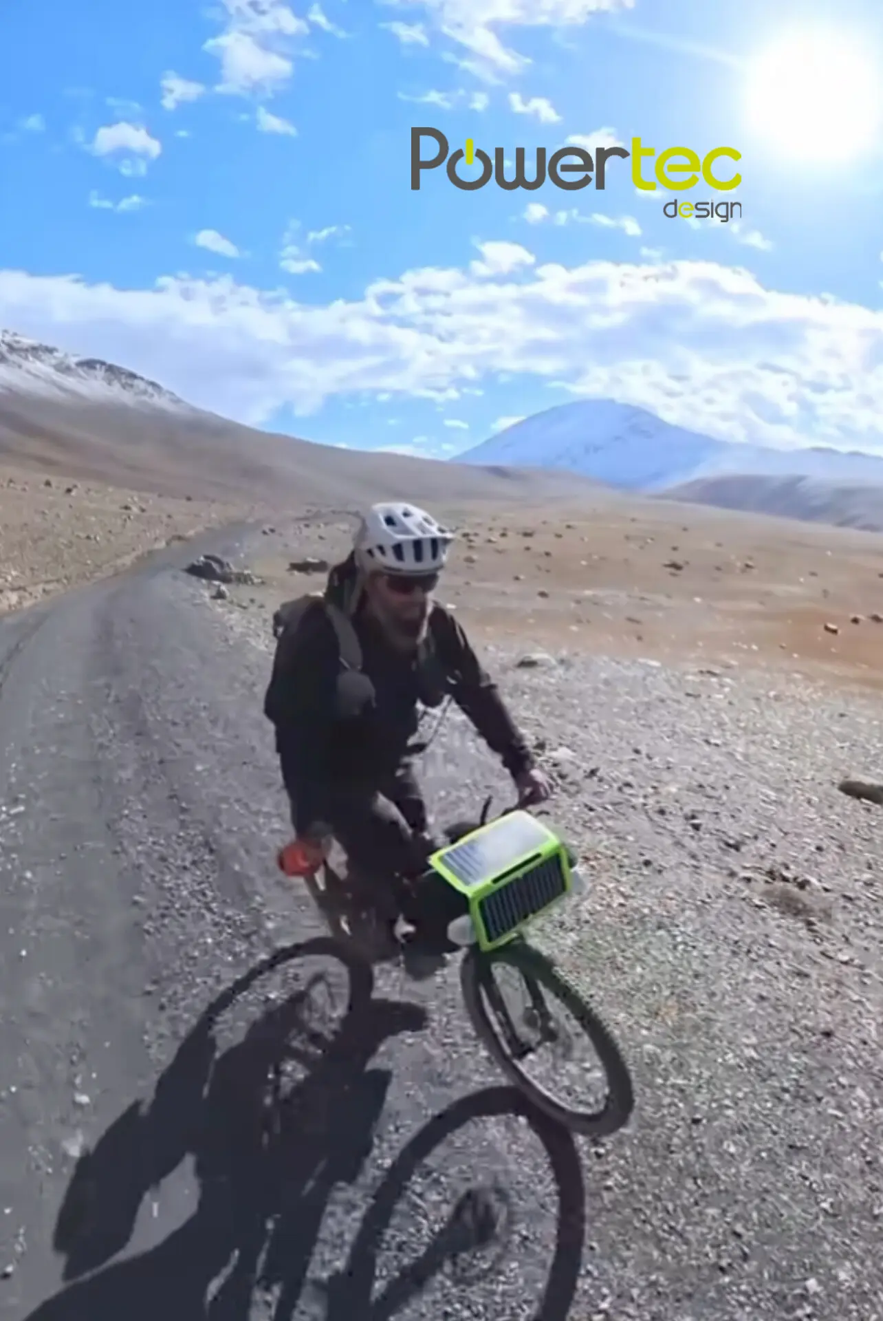 Cycliste équipé d’un casque et d’un sac à dos sur une route poussiéreuse en montagne, avec une batterie/panneau solaire portable vert fosforescent fixé au guidon.